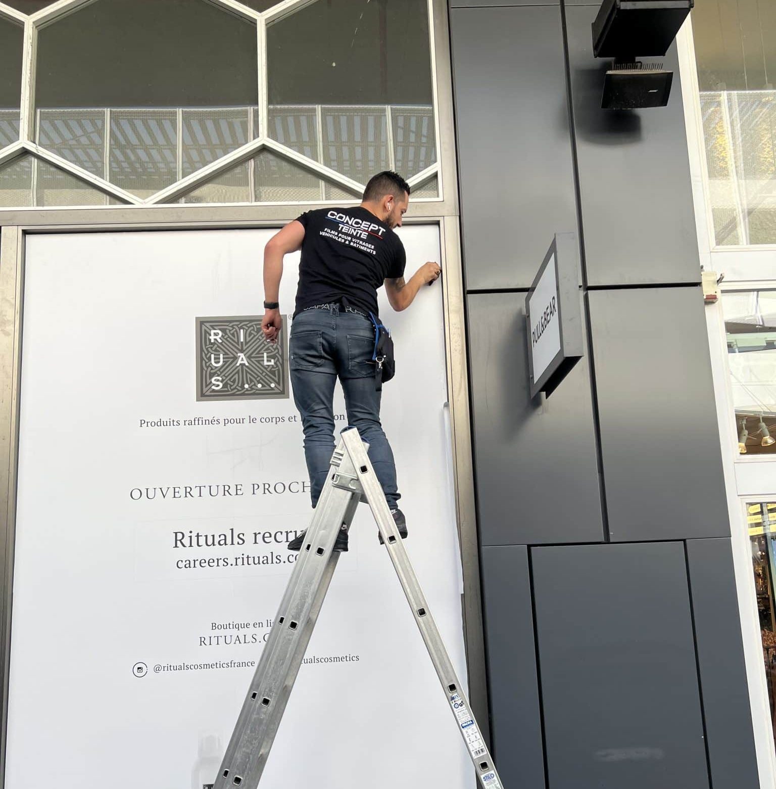 Technicien Concept Teinte sur un escabeau posant un grand visuel imprimé sur la vitrine d'une boutique en travaux